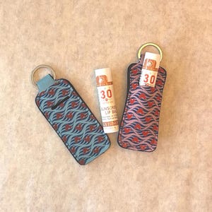 May include: Two lip balm holders with key rings, one blue and one pink, each with a repeating wave and heart pattern. A tube of sunscreen lip balm is next to the holders. The lip balm has the number 30 printed on it.