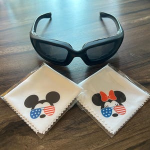 May include: Black sunglasses with a thick frame rest above two white cleaning cloths. Each cloth features a cartoon mouse head wearing sunglasses with red, white, and blue stripes and stars. One has a red bow.