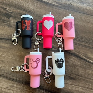 May include: A collection of six colorful tumbler keychains. The keychains are black, pink, and white, with various designs including hearts, Mickey Mouse, and the word "LOVE". Each tumbler has a handle and a straw.