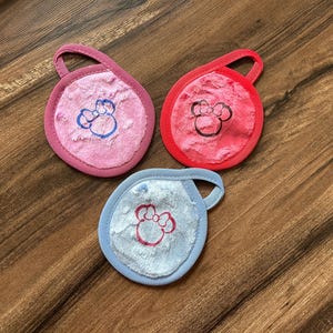 May include: Three soft, round, fabric face cloths with a bow design. The cloths are pink, red, and blue.