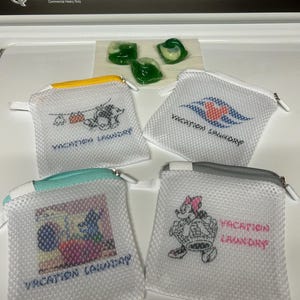 May include: Four white mesh laundry bags with zippers, each featuring unique character graphics and the text "VACATION LAUNDRY". The bags have yellow, white, teal, and gray zipper accents, and are displayed with three green laundry detergent pods.
