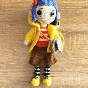 May include: A crocheted doll with blue hair, wearing a yellow raincoat, brown skirt, and black and white striped socks. The doll is holding a brown bag.