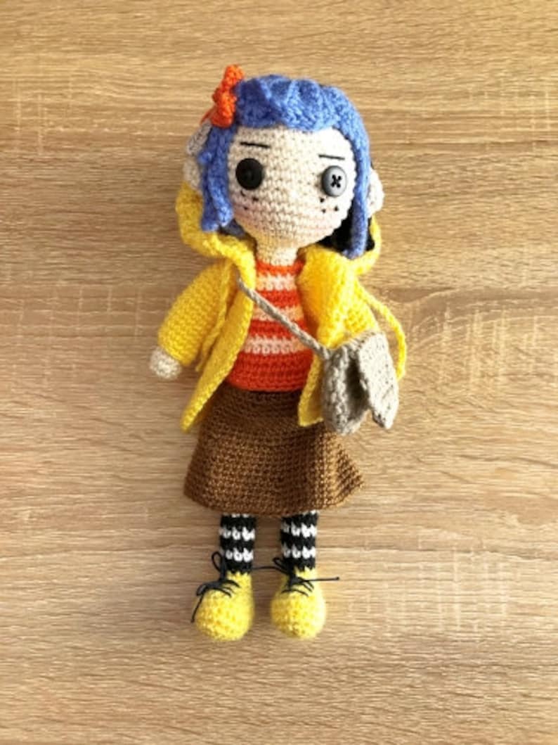 May include: A crocheted doll with blue hair, wearing a yellow raincoat, brown skirt, and black and white striped socks. The doll is holding a brown bag.