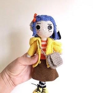May include: A crocheted doll with blue hair, a yellow jacket, a brown skirt, and a gray bag. The doll is wearing black and white striped socks and yellow shoes.