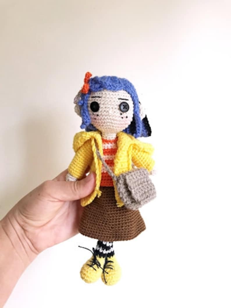 May include: A crocheted doll with blue hair, a yellow jacket, a brown skirt, and a gray bag. The doll is wearing black and white striped socks and yellow shoes.