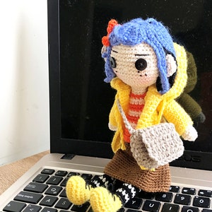 May include: Crocheted doll with blue hair, wearing a yellow raincoat, orange and white striped shirt, brown skirt, and grey bag. The doll is sitting on a laptop keyboard.