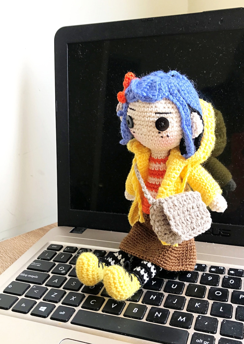 May include: Crocheted doll with blue hair, wearing a yellow raincoat, orange and white striped shirt, brown skirt, and grey bag. The doll is sitting on a laptop keyboard.