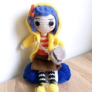 May include: A crocheted doll wearing a yellow raincoat, blue hair, and a striped shirt. The doll is holding a small gray bag and sitting on a blue yarn ball.