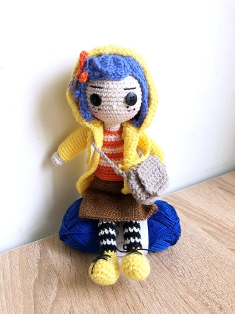 May include: A crocheted doll wearing a yellow raincoat, blue hair, and a striped shirt. The doll is holding a small gray bag and sitting on a blue yarn ball.