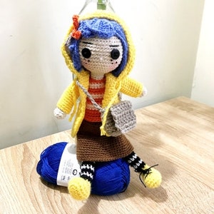 May include: Crocheted doll wearing a yellow raincoat, blue hair, and a brown skirt. The doll is holding a gray crocheted bag and is sitting on a blue yarn ball.