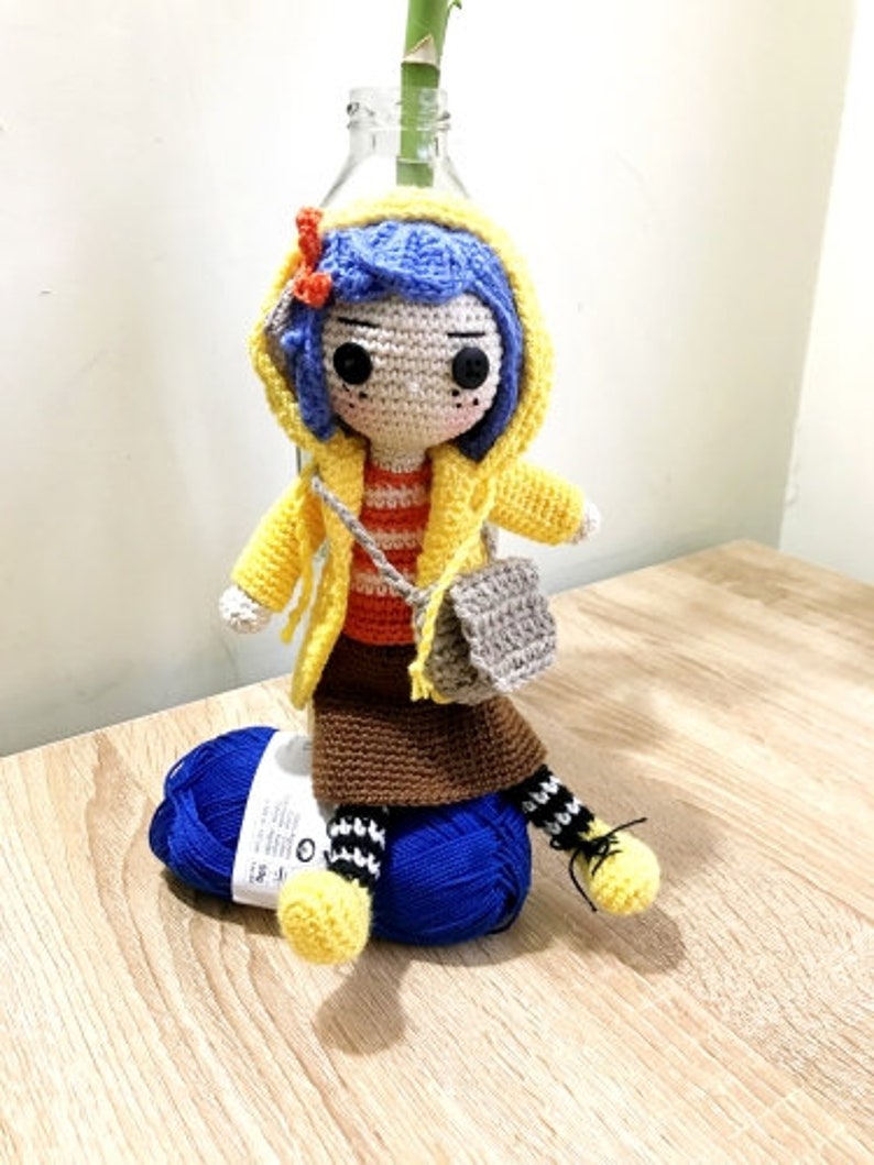 May include: Crocheted doll wearing a yellow raincoat, blue hair, and a brown skirt. The doll is holding a gray crocheted bag and is sitting on a blue yarn ball.