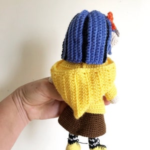 May include: A crocheted doll with blue hair, a yellow sweater, a brown skirt, and black and white striped socks. The doll is wearing yellow shoes.
