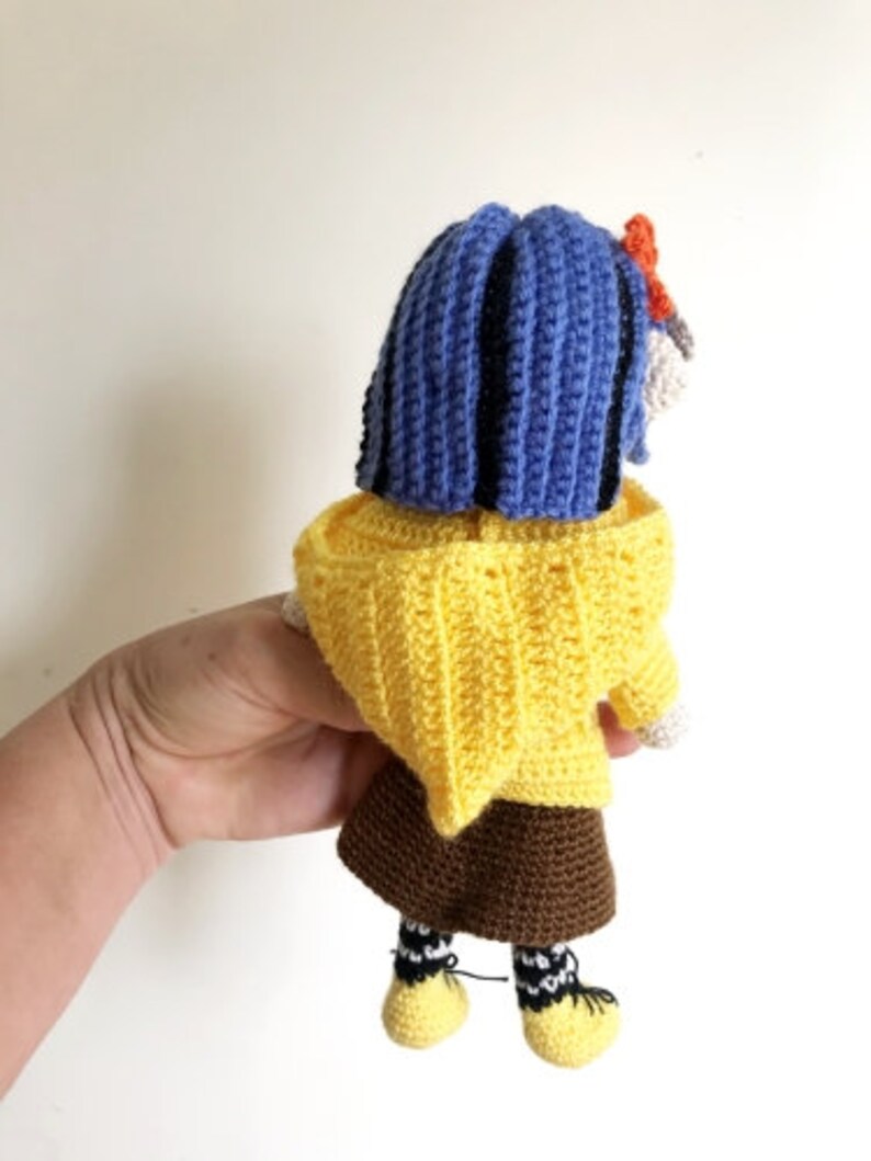 May include: A crocheted doll with blue hair, a yellow sweater, a brown skirt, and black and white striped socks. The doll is wearing yellow shoes.
