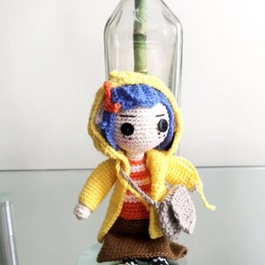 May include: A crocheted doll wearing a yellow raincoat, a brown skirt, and a striped shirt. The doll has blue hair and brown eyes. The doll is holding a small gray bag.