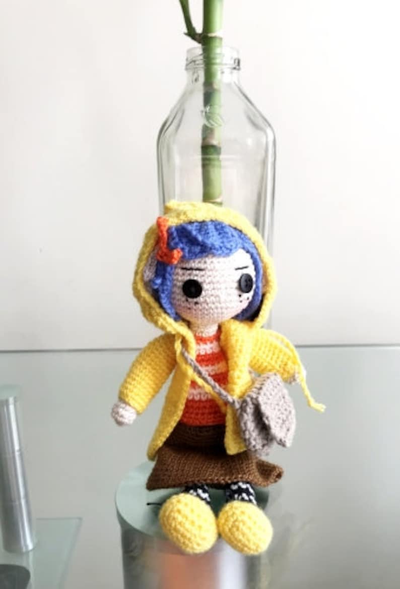 May include: A crocheted doll wearing a yellow raincoat, a brown skirt, and a striped shirt. The doll has blue hair and brown eyes. The doll is holding a small gray bag.