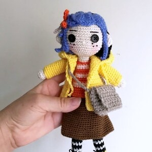 May include: Crocheted doll with blue hair, wearing a yellow raincoat, brown skirt, and black and white striped socks. The doll is holding a gray crocheted bag.