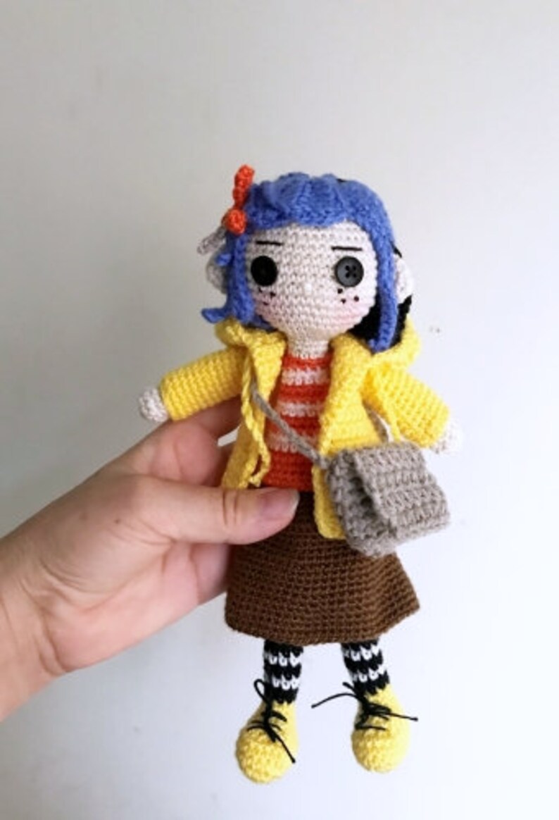 May include: Crocheted doll with blue hair, wearing a yellow raincoat, brown skirt, and black and white striped socks. The doll is holding a gray crocheted bag.