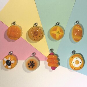May include: A collection of eight miniature food-themed charms featuring various baked goods and snacks, including a melon pan, a donut with sprinkles, a round bun, a baguette, a pastry with a dark center, a sesame seed bun, a spiral snack, and an egg bun.