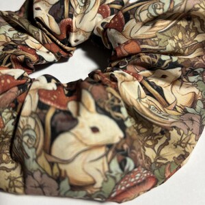 Puede incluir: Un scrunchie de tela marr&oacute;n, blanco y verde con un patr&oacute;n repetido de conejos blancos, setas y hojas.