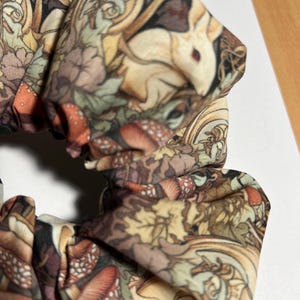 Puede incluir: Un scrunchie de tela marr&oacute;n, blanco y verde con un estampado de bosque caprichoso que presenta hongos, hojas y un conejo blanco.
