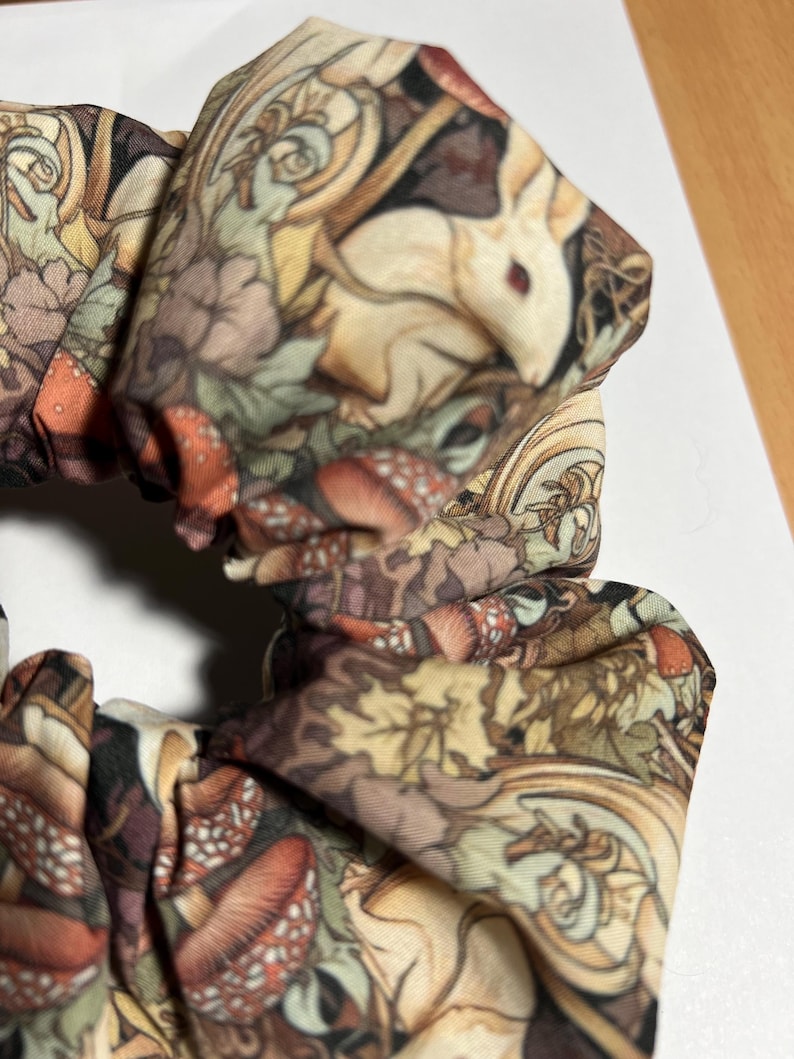 Puede incluir: Un scrunchie de tela marr&oacute;n, blanco y verde con un estampado de bosque caprichoso que presenta hongos, hojas y un conejo blanco.