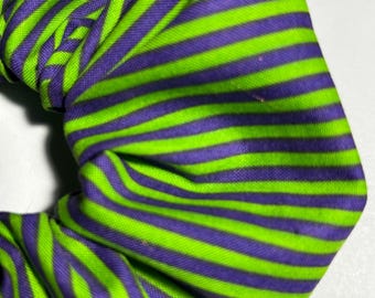 May include: Close-up of a scrunchie with a repeating pattern of green and purple horizontal stripes. The fabric is gathered and twisted, creating a textured appearance. The scrunchie is likely used to hold hair in a ponytail or bun.