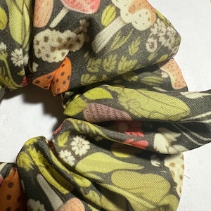 May include: A fabric scrunchie featuring a dark background with a repeating pattern of mushrooms, leaves, and flowers in pink, orange, white, and green. This is a hair accessory.