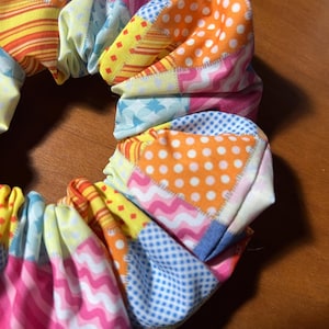 May include: A vibrant scrunchie with a patchwork design. The fabric incorporates diverse patterns, such as polka dots, stripes, and geometric shapes, in shades of pink, orange, yellow, and blue. The scrunchie is gathered and elasticated.