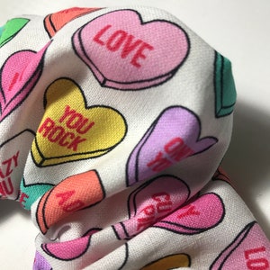 May include: White fabric with a repeating pattern of colourful candy hearts. The hearts have different messages on them, including "LOVE", "YOU ROCK", and "CRAZY FOR YOU".