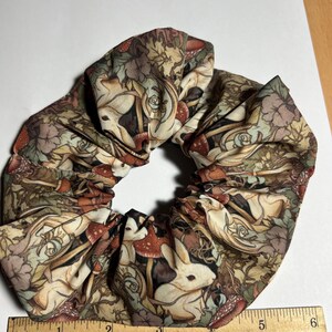 Puede incluir: Un scrunchie de tela marr&oacute;n, beige y blanco con un patr&oacute;n repetido de conejos blancos, setas y hojas. El scrunchie tiene un di&aacute;metro de aproximadamente 10 cm.