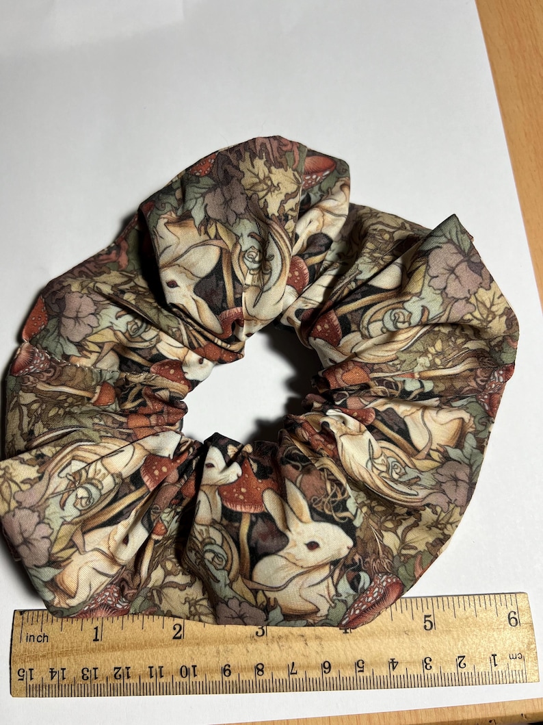 Puede incluir: Un scrunchie de tela marr&oacute;n, beige y blanco con un patr&oacute;n repetido de conejos blancos, setas y hojas. El scrunchie tiene un di&aacute;metro de aproximadamente 10 cm.
