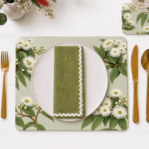 May include: A rectangular placemat featuring a botanical illustration of white eucalyptus flowers and green leaves, a white dinner plate, a solid olive green napkin with a white scalloped edge, a gold fork, a gold knife, and a gold spoon.