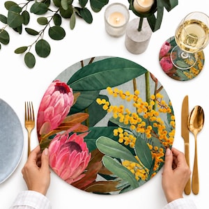 May include: A round placemat featuring a botanical illustration of two large pink protea flowers and clusters of bright yellow mimosa blossoms set against dark green leaves. The design is printed on a circular surface with a smooth finish.
