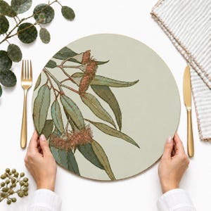 May include: A round placemat featuring a light sage green background with a detailed botanical illustration of eucalyptus branches. The design includes elongated green leaves and textured, reddish-brown eucalyptus flower buds.