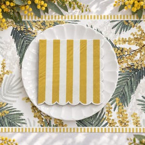 May include: A white scalloped edge paper napkin with vertical mustard yellow and white stripes, placed on a white plate. The plate rests on a placemat featuring a pattern of green palm leaves and yellow mimosa flowers.