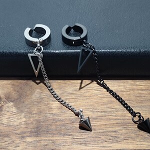 May include: Two non-piercing ear cuffs. One is silver-toned, the other black. Each cuff has a dangling chain with a triangle pendant and a pointed end. The cuffs are displayed on a dark surface with a wooden background.