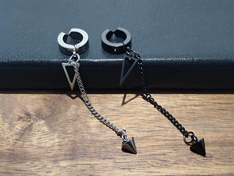 May include: Two non-piercing ear cuffs. One is silver-toned, the other black. Each cuff has a dangling chain with a triangle pendant and a pointed end. The cuffs are displayed on a dark surface with a wooden background.