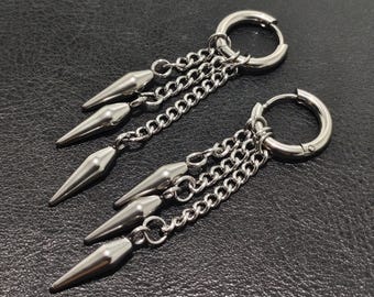 May include: A pair of silver-colored hoop earrings with dangling chains and pointed cone-shaped charms. The earrings are made of a shiny metal and are displayed on a dark surface.