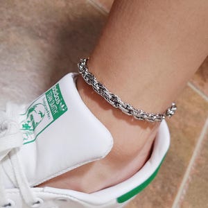 May include: A silver chain ankle bracelet with a twisted rope design. The bracelet is worn on a person's ankle, and the person is wearing white sneakers with green accents and the text "adidas Stan Smith" on the side.