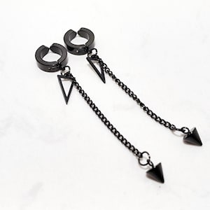 May include: A pair of black clip-on earrings. Each earring features a circular clip, a triangular pendant, a chain, and a cone-shaped charm. The earrings are displayed on a white surface.