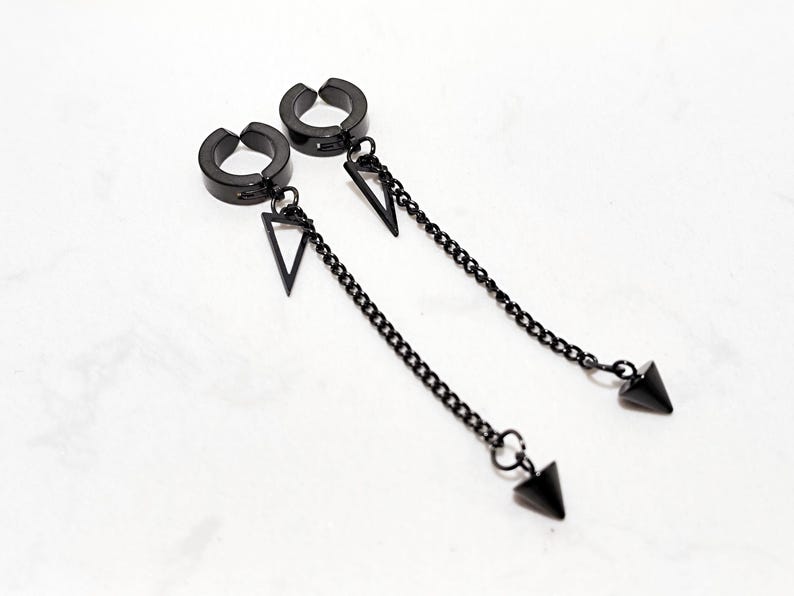 May include: A pair of black clip-on earrings. Each earring features a circular clip, a triangular pendant, a chain, and a cone-shaped charm. The earrings are displayed on a white surface.