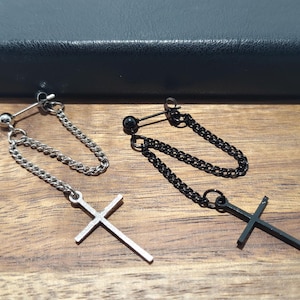 May include: Two pairs of dangle earrings. One pair is silver-toned with a cross pendant and chain. The other pair is black with a cross pendant and chain. The earrings are displayed on a wooden surface.