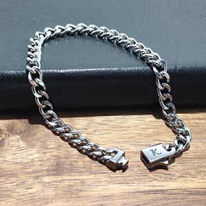 May include: A silver curb chain bracelet with a clasp that has the letter "K" engraved on it.
