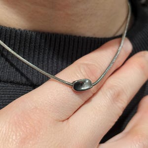 May include: A silver necklace featuring a small, oval pendant with a delicate design. The necklace is worn around the neck. The image is a close-up of the necklace and the person wearing it.