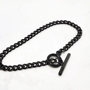 May include: A black chain bracelet with a toggle clasp. The bracelet features a series of small, rounded links. The toggle clasp is a simple design, with a circular ring and a straight bar. The bracelet is made of a dark, shiny metal.