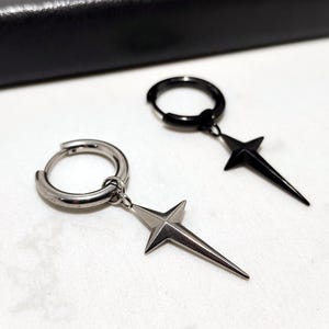 May include: Two hoop earrings with star-shaped pendants. One earring is silver-toned, the other is black. The star pendants have a pointed design. The earrings are displayed on a white surface.