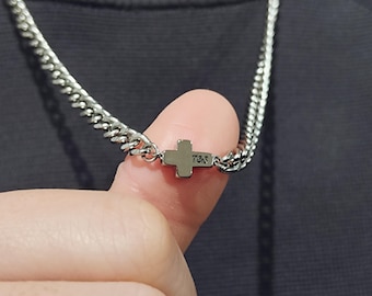 May include: A silver chain necklace with a small silver cross pendant. The cross has the inscription "7.5S" on the back.