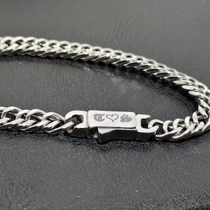 May include: A silver curb chain necklace with a rectangular clasp. The clasp has engraved symbols and a heart. The chain is made of interlocking links and is displayed on a dark surface.