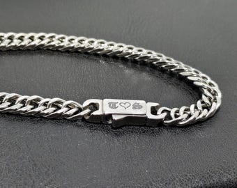 May include: A silver-colored curb chain necklace with a rectangular clasp. The clasp features engraved symbols and a heart. The chain is made of interlocking links and is displayed on a dark surface.