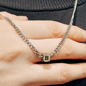 May include: A silver chain necklace with a small silver cube pendant featuring the letter "B" in black.